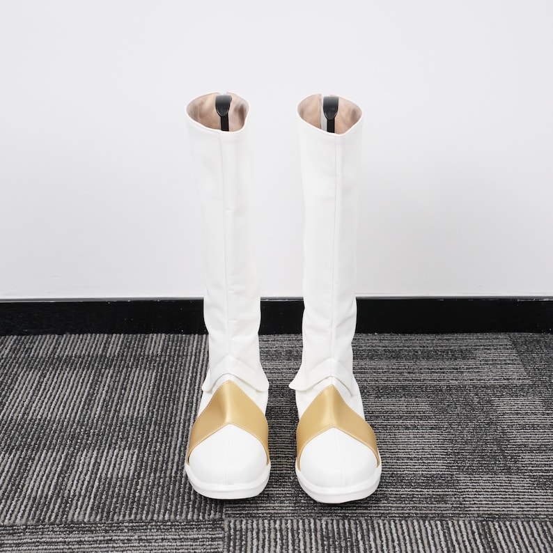 May include: A pair of white knee-high boots with gold accents. The boots have a smooth, clean design with a gold band across the top of the foot. The interior of the boots is a light tan color.