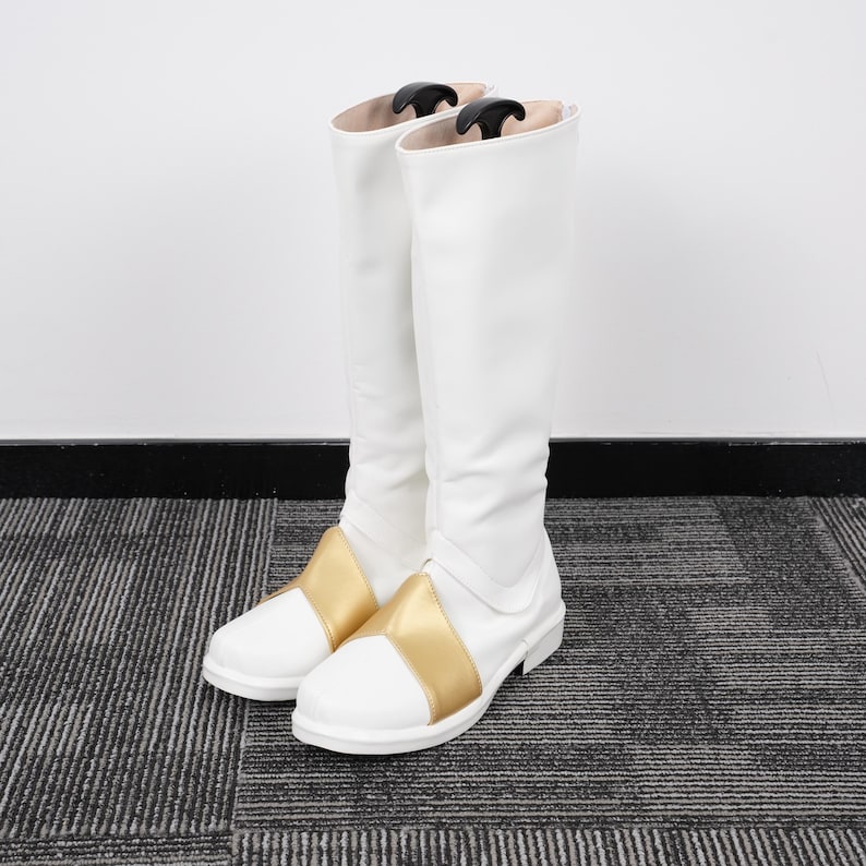 May include: A pair of white knee-high boots with a gold band across the top of the foot. The boots have a smooth, clean design and are standing upright. The boots are made of a shiny material and are likely for costume or fashion purposes.