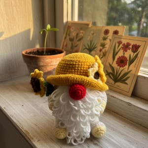 以下が含まれることがあります： 黄色いヒマワリの帽子、赤い鼻、白いひげの、手作りのかぎ針編みのノーム。ノームは小さなヒマワリを持っています。ノームは、背景にヴィンテージの花柄プリントのある白い窓枠に座っています。