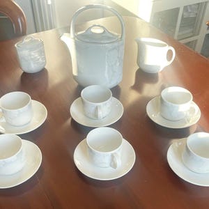 Könnte beinhalten: Ein weißes Porzellan-Teeservice mit Teekanne, Sahnekännchen, Zuckerdose und sechs Tassen mit Untertassen. Das Set steht auf einem Holztisch. Das Teeservice hat ein dezentes Blumenmuster.