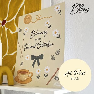 May include: A beige art print with the text "Blooming with Tea and Stitches." The print features illustrations of yarn, daisies, a teacup, a bow, and knitting needles. The print is labeled "Art Print in A3."