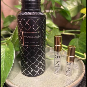 May include: A black perfume bottle with a silver geometric pattern and the text "VANGUARD ROYAL COLLECTION" is displayed on a ceramic plate. Two small sample bottles with gold caps are next to the perfume bottle. Green leaves are in the background.