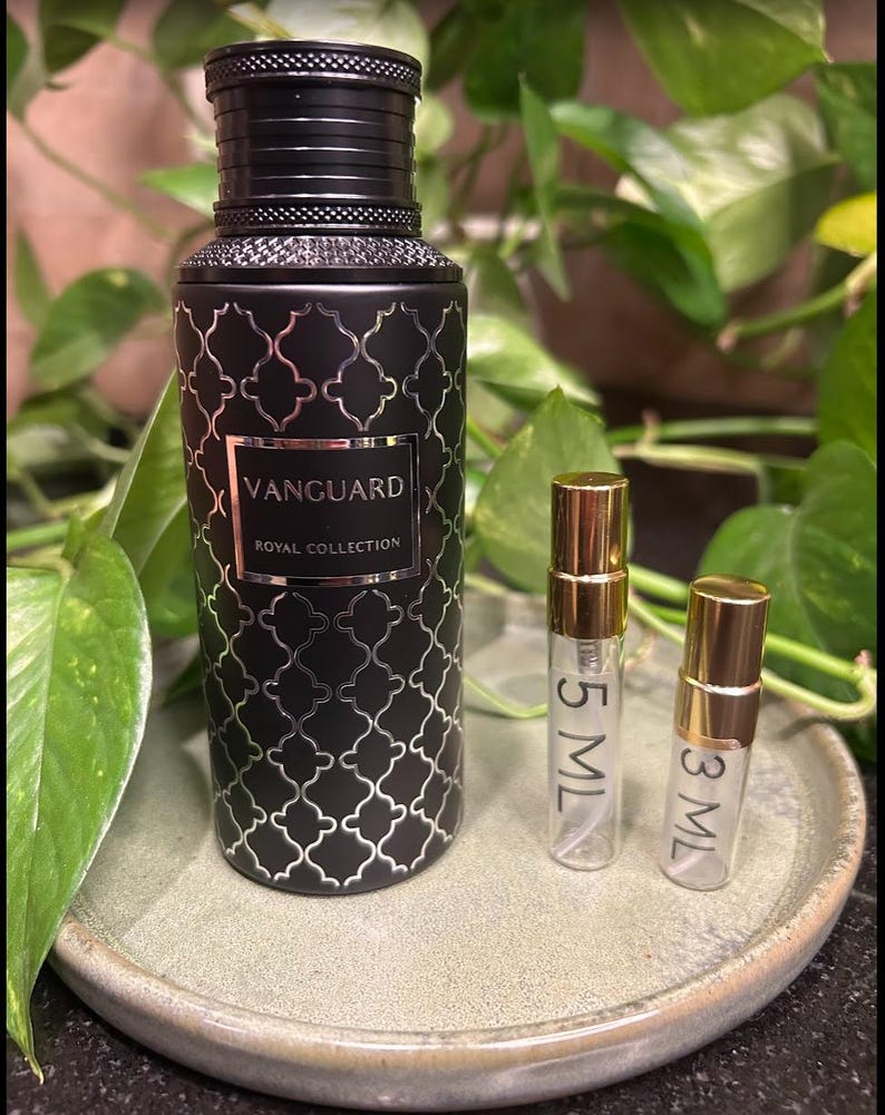May include: A black perfume bottle with a silver geometric pattern and the text "VANGUARD ROYAL COLLECTION" is displayed on a ceramic plate. Two small sample bottles with gold caps are next to the perfume bottle. Green leaves are in the background.