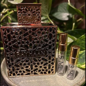 Puede incluir: Un frasco de perfume dorado con intrincados cortes geom&eacute;tricos, la palabra "RAYHAAN" es visible en la parte delantera. Dos peque&ntilde;os frascos de vidrio con tapas doradas est&aacute;n junto al frasco, etiquetados 5 ML y 3 ML. Los art&iacute;culos est&aacute;n sobre un plato gris y redondo.
