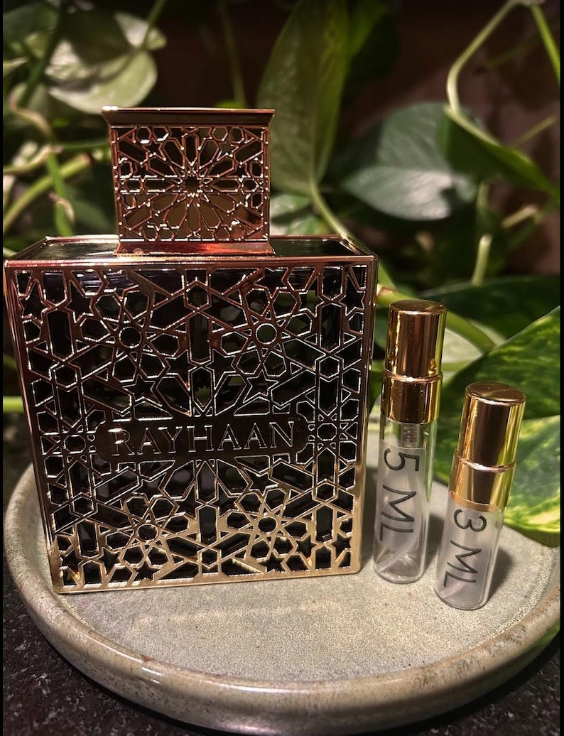 Puede incluir: Un frasco de perfume dorado con intrincados cortes geom&eacute;tricos, la palabra "RAYHAAN" es visible en la parte delantera. Dos peque&ntilde;os frascos de vidrio con tapas doradas est&aacute;n junto al frasco, etiquetados 5 ML y 3 ML. Los art&iacute;culos est&aacute;n sobre un plato gris y redondo.