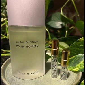 May include: A frosted glass bottle of L'Eau d'Issey Pour Homme cologne with a light gray cap, alongside two small sample vials. The vials are labeled 5 ML and 3 ML, with gold-colored caps. The items rest on a green ceramic plate.