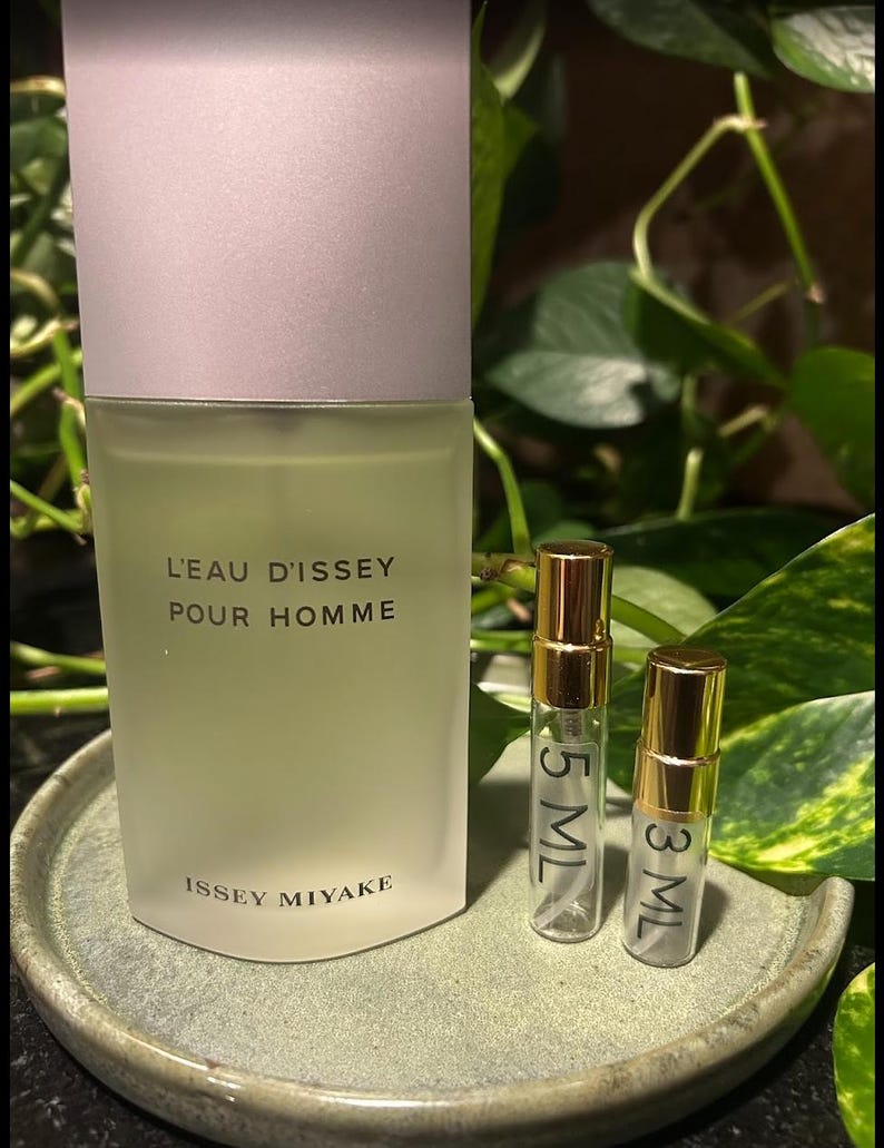 May include: A frosted glass bottle of L'Eau d'Issey Pour Homme cologne with a light gray cap, alongside two small sample vials. The vials are labeled 5 ML and 3 ML, with gold-colored caps. The items rest on a green ceramic plate.