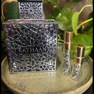 May include: A silver perfume bottle with intricate geometric cutouts and the name "RAYHAAN" etched on the front. Two small sample bottles with gold caps are next to the perfume bottle. The bottles are on a green ceramic plate.