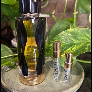May include: A perfume bottle with a black cap and gold accents, featuring a decorative snake design. The bottle is filled with a golden liquid and is accompanied by two smaller sample bottles labeled 5 ML and 3 ML.