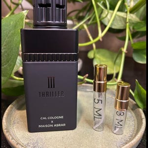 Pode incluir: Um frasco de col&ocirc;nia preto com uma tampa texturizada e a palavra "Thriller" impressa na frente. Dois pequenos frascos de amostra com tampas douradas est&atilde;o ao lado do frasco. A col&ocirc;nia &eacute; da Cal Cologne x Maison Asrar.
