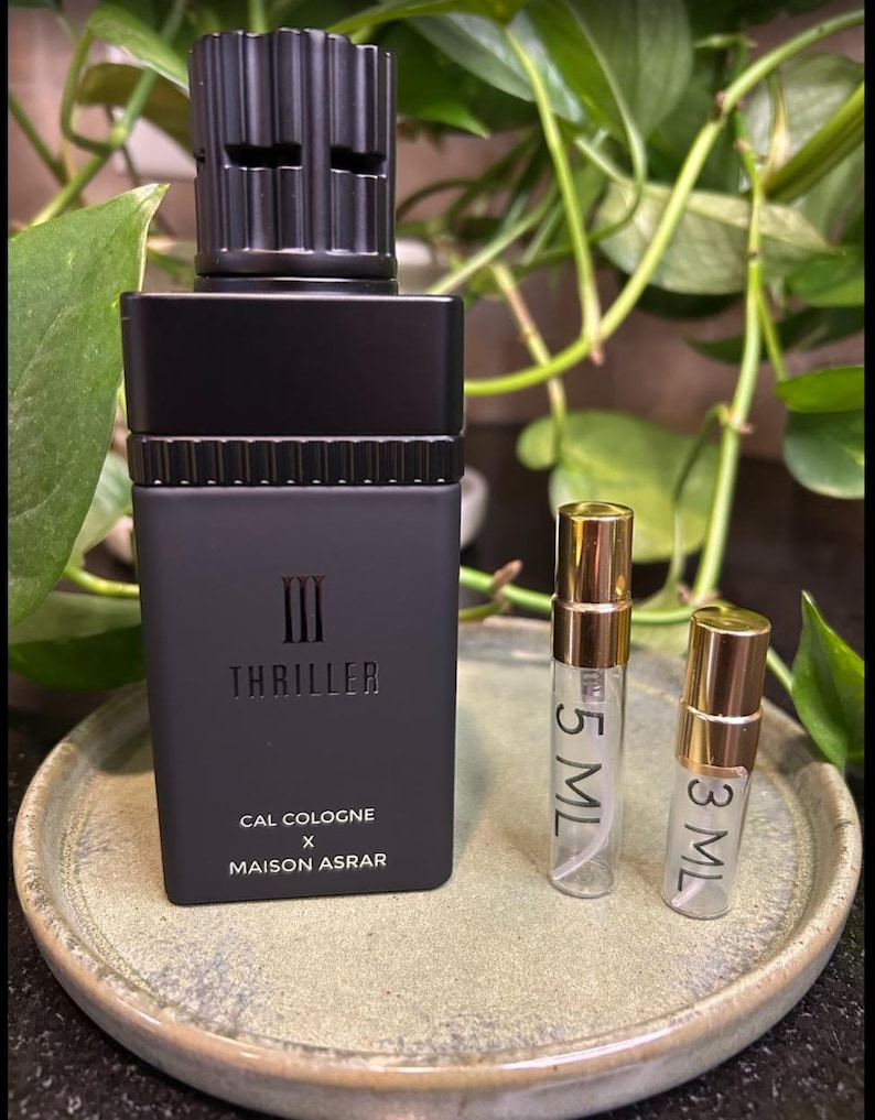Pode incluir: Um frasco de col&ocirc;nia preto com uma tampa texturizada e a palavra "Thriller" impressa na frente. Dois pequenos frascos de amostra com tampas douradas est&atilde;o ao lado do frasco. A col&ocirc;nia &eacute; da Cal Cologne x Maison Asrar.