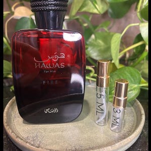 May include: A red and black perfume bottle with the text "HAWAS For Him FIRE" and two small sample bottles on a green plate. The perfume bottle has a black cap with a textured design. The sample bottles are labeled "5 ML" and "3 ML".