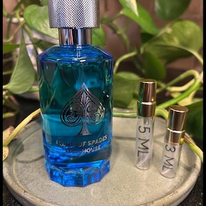 May include: A blue perfume bottle with a silver cap and the words "GAME OF SPADES FULL HOUSE" printed on the front. The bottle has a spade symbol. Two small sample bottles are next to the perfume bottle. The bottles are on a round, gray plate.