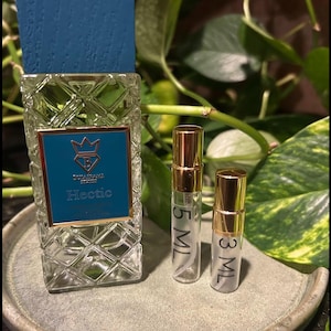 May include: A clear glass perfume bottle with a teal label and a blue cap, labeled "Hectic." Two smaller sample bottles with gold caps are also present. The bottles sit on a gray ceramic tray, with green foliage in the background.