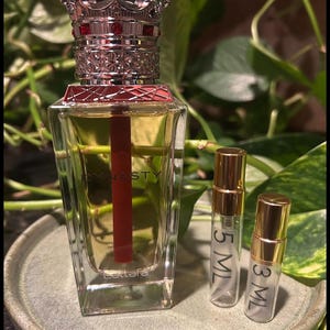 Puede incluir: Un frasco de perfume con tap&oacute;n en forma de corona y un acento rojo, etiquetado "Dynasty" y "Lattafa". Tambi&eacute;n hay dos frascos m&aacute;s peque&ntilde;os, etiquetados "5 ML" y "3 ML". Los frascos est&aacute;n sobre un plato redondo y gris, con follaje verde de fondo.