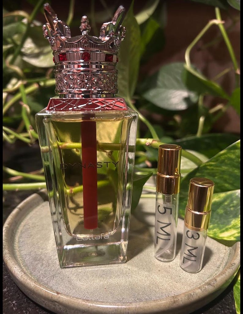 Puede incluir: Un frasco de perfume con tap&oacute;n en forma de corona y un acento rojo, etiquetado "Dynasty" y "Lattafa". Tambi&eacute;n hay dos frascos m&aacute;s peque&ntilde;os, etiquetados "5 ML" y "3 ML". Los frascos est&aacute;n sobre un plato redondo y gris, con follaje verde de fondo.