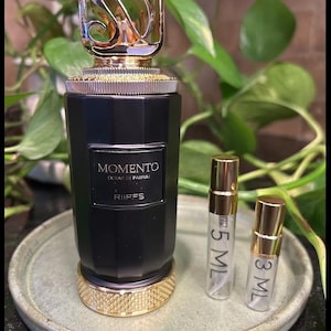 May include: A black perfume bottle with a gold-colored cap and base, labeled "Momento Extrait de Parfum Riffs." Two small sample vials, one 5 ml and one 3 ml, are next to the bottle. The bottle sits on a light green plate.