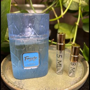 May include: A light blue perfume bottle with the word "Freeze" on the front, along with two small perfume vials. The vials are labeled "5 ML" and "3 ML" and have gold caps. The items are on a round, light green plate.