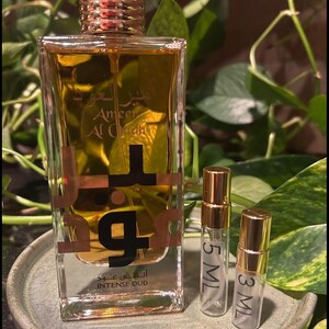 May include: A clear glass perfume bottle with a gold cap and a label that reads "Ameer Al Oudh Intense Oud." Two smaller sample bottles, labeled 5 ML and 3 ML, are also present. The bottles sit on a round, light green tray.