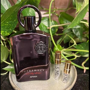 May include: A dark purple perfume bottle with a silver cap and a decorative emblem. The bottle is labeled "SUPREMACY not only intense AFNAN." Two small sample vials with gold caps are next to the bottle. The background features green foliage.