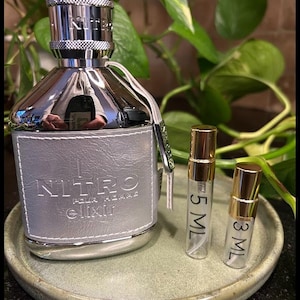 May include: A silver-toned perfume bottle with a gray leather-like label, embossed with "NITRO pour homme elixir." Two small sample vials, labeled 5 ML and 3 ML, sit beside it on a green ceramic tray. The bottle has a silver cap.