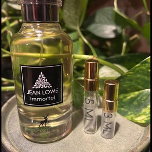 May include: A perfume bottle with a black label that reads "JEAN LOWE immortel" and a deer logo. Two smaller sample bottles with gold caps are next to the larger bottle. The bottles are on a gray ceramic plate.