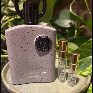 May include: A perfume bottle with a speckled gray finish, a dark brown cap, and a circular handle. The bottle is labeled "SUPREMACY COLLECTOR'S EDITION AFNAN." Two small glass vials with gold caps, labeled 5 ML and 3 ML, sit beside it.