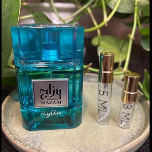 May include: A turquoise perfume bottle with a matching cap, a silver label with Arabic script and the word "MAZAAJ", and the word "rhythm" in cursive. Two small sample vials with gold caps are next to the bottle. The vials are labeled "5 ML" and "3 ML".