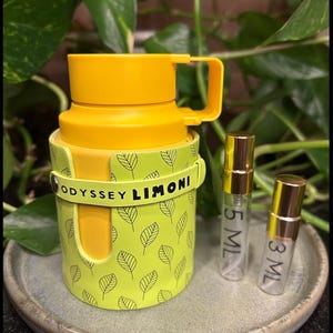 May include: A yellow travel mug with a handle and a light green sleeve featuring a leaf pattern. The sleeve has the text "ODYSSEY LIMONI". Two small perfume bottles are next to the mug, one labeled "5 ML" and the other "3 ML".