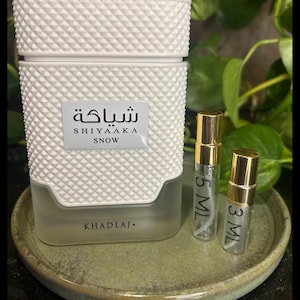 May include: A white perfume bottle with a textured diamond pattern, labeled "SHIYAAKA SNOW" in English and Arabic. Two small sample bottles with gold caps are next to the perfume bottle. The bottles are on a green ceramic plate.