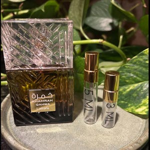 Puede incluir: Un frasco de perfume con una tapa transparente con dise&ntilde;o geom&eacute;trico y una base marr&oacute;n oscuro. El frasco lleva la inscripci&oacute;n "Khamrah Qahwa Lattafa" en letras doradas y escritura &aacute;rabe. Tambi&eacute;n hay dos peque&ntilde;os frascos pulverizadores transparentes con tapas doradas, etiquetados con 5 ml y 3 ml.