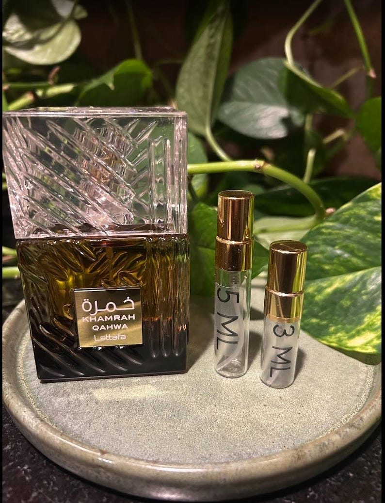 Puede incluir: Un frasco de perfume con una tapa transparente con dise&ntilde;o geom&eacute;trico y una base marr&oacute;n oscuro. El frasco lleva la inscripci&oacute;n "Khamrah Qahwa Lattafa" en letras doradas y escritura &aacute;rabe. Tambi&eacute;n hay dos peque&ntilde;os frascos pulverizadores transparentes con tapas doradas, etiquetados con 5 ml y 3 ml.