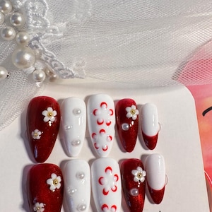Könnte beinhalten: Ein Set von Press-On-Nägeln mit einem rot-weißen Blumendesign. Die Nägel haben eine tiefrote Glitzerbasis mit weißen Blumenakzenten und Perlen. Einige Nägel sind weiß mit roten Blumenmustern.