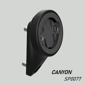 Puede incluir: Soporte negro para ciclocomputador con diseño circular y dos postes plateados. El soporte tiene el texto "CANYON SP0077" impreso. El soporte está diseñado para sujetar un ciclocomputador.