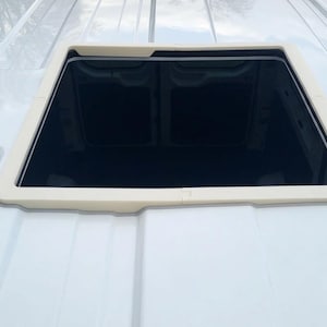 May include: A rectangular, black-tinted skylight with a cream-coloured frame is set into the white roof of a vehicle. The skylight is open, revealing a view of the sky. The roof has a textured surface.