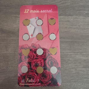 May include: A pink card with the text "12 mois secret" and a floral design. The card features gold heart and circle-shaped scratch-off areas, and a graphic of a white envelope. The bottom of the card has a rose graphic.