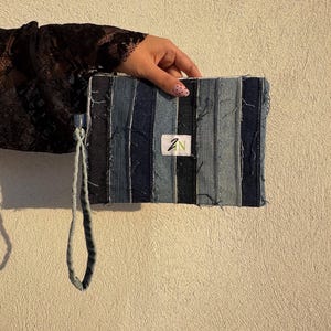 Può includere: Pochette rettangolare in denim con strisce verticali di varie tonalità di blu. La pochette ha un cinturino da polso e una piccola etichetta bianca con le lettere "ZN" in verde. I bordi del denim appaiono sfilacciati.