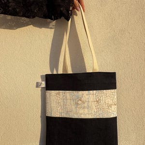 May include: Black tote bag with a cream-colored strap. The bag features a horizontal band with a textured, off-white pattern. The bag is held by a person wearing a black lace sleeve.