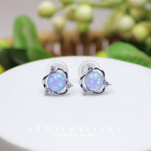 May include: A pair of stud earrings featuring a light blue opal stone set in a silver-tone floral design. Each earring has a round opal center and three small clear accent stones. The text "ARWJEWELLERY Handcrafted Jeweller" is at the bottom.