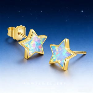 May include: A pair of gold-colored star-shaped stud earrings. Each earring features a five-pointed star with a colorful, iridescent opal-like center. The earrings are set against a gradient blue background with small white stars.