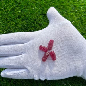 May include: Four small, raw, ruby gemstones are displayed on a white glove against a backdrop of green grass. The rubies are a deep red color and appear to be uncut, with a natural, rough texture. The gemstones are arranged in a star-like pattern.