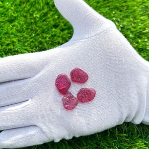 Può includere: Quattro piccole pietre preziose rosse, irregolari e traslucide con una superficie sfaccettata e ruvida. Le pietre sono appoggiate su una superficie coperta da un tessuto bianco, mettendo in risalto il loro colore cremisi e l'aspetto naturale.