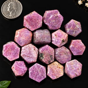 Könnte beinhalten: Eine Ansammlung roher, sechseckiger rosa Rubinkristalle mit texturierten Oberflächen in verschiedenen Magenta- und Violetttönen. Eine silberfarbene US-Vierteldollarmünze ist zur Größenorientierung in der oberen linken Ecke abgebildet.