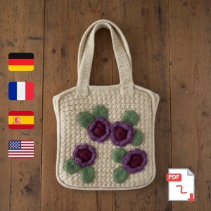 Könnte beinhalten: Beige gehäkelte Tragetasche mit quadratischer Form und zwei Griffen. Die Tasche ist mit drei lila Blumenapplikationen mit grünen Blättern verziert. Die Tasche steht auf einer Holzoberfläche.