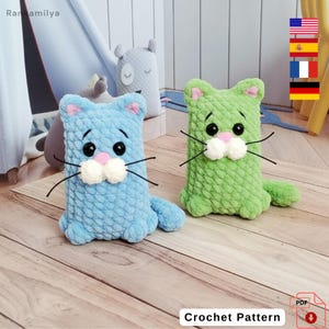 May include: Two crochet cat toys, one light blue and one green, sit side-by-side on a wooden surface. Each cat has black eyes, pink accents, and white whiskers. The text "Crochet Pattern" is visible at the bottom of the image.