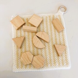 May include: A collection of wooden geometric blocks, including squares, triangles, and a half-circle, are arranged on a yellow and white crocheted mat. A wooden ring is attached to the mat. The blocks are made of light-coloured wood.