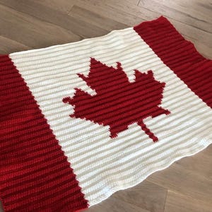 May include: A red, white, and red crocheted blanket featuring the Canadian flag design. The blanket has a red maple leaf in the centre, with red stripes on the top and bottom, and white stripes in the middle. The blanket is lying on a wooden surface.
