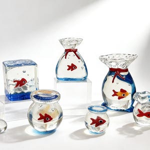 Peut inclure: Plusieurs sculptures en verre transparent, chacune contenant un poisson rouge. Les sculptures ont des formes variées, comme des cubes, des sacs et des bols. Les bases sont bleues, et certaines ont des détails rouges.