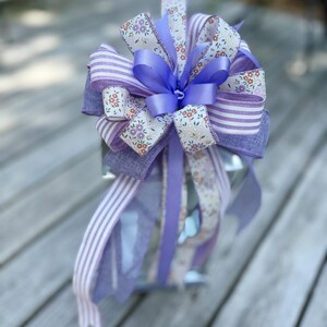 May include: A decorative bow crafted from multiple ribbons in shades of lavender and purple. The design features a mix of solid purple satin, lavender and white striped fabric, and a cream-colored ribbon with a small floral print pattern.