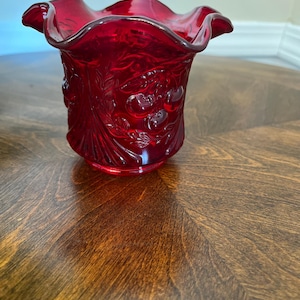 May include: A ruby red glass votive candle holder with a ruffled rim. The sides of the holder are decorated with a relief pattern of cherries and leaves. The candle holder sits on a dark wooden surface.